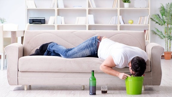JUnger Mann liegt auf einer Couch und beugt sich über einen davor stehenden Eimer. Daneben steht eine Flasche und ein Glas.