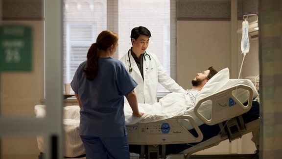 Ein Arzt steht an einem Krankenbett und spricht mit einem Patienten.
