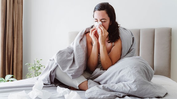 Eine junge Frau sitzt im Bett und putzt sich die Nase