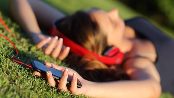 Mädchen liegt mit Kopfhörern im Gras und hält Smartphone