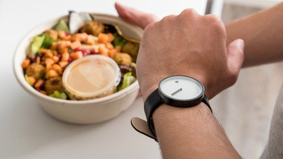 Symbolbild: Ein junger Mann schaut auf seine Armbanduhr, während er nach einem Salat mit Falafel, Hummus, Süßkartoffeln, Roter Bete und Granatapfelkernen greift.