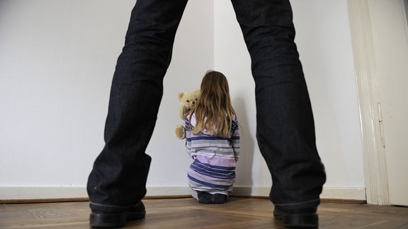 Durch die breit aufgestellten Beine einer Person, sieht man ein Kind mit einem Kuscheltier im Arm mit Blickrichtung zur Wand auf dem Boden sitzen.