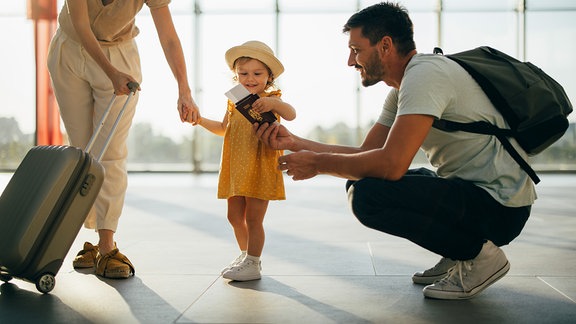 Fröhlich lächelnder Ehemann und seine Frau stehen mit Gepäck und ihr kleines Mädchen mit Dokumenten in den Händen am Flughafen
