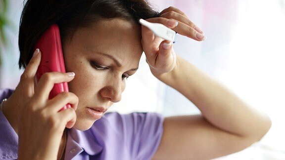 Eine Frau mit einem Fieberthermometer inder Hand, die einen Anruf tätigt.