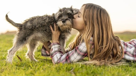 Ein Mädchen liegt auf einer Wiese und küsst ihren Hund.