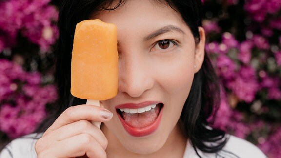 Junge fröhliche Frau bedeckt eins ihrer Augen mit einem orangenem Eis am Stiel.