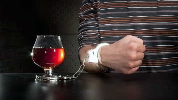 Symbolbild - Hand mit Handschelle an einem Glas Alkohol.