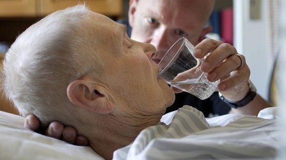 Seniorin wird dabei geholfen aus einem Wasserglas zu trinken.