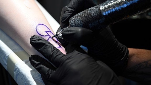 Eine Tattoo-Künstlerin tätowiert zwei Halbkreise und einen ganzen Kreis auf einen Unterarm. Das Motiv wurde für eine Aktion des Münchner Vereins Junge Helden entworfen und soll ein Statement zum Thema Organspenden sein
