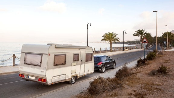 Pkw mit Wohnwagen fährt auf einer Straße am Meer