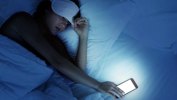 Frau mit Handy unnd Schlafbrille im Bett