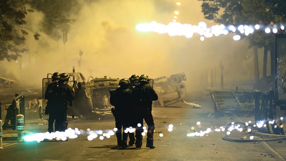 Beamte der Bereitschaftspolizei patrouillieren bei Unruhen auf einer Straße, während Feuerwerkskörper explodieren. 