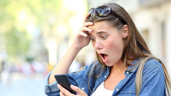 Schockierte junge Frau schaut aufs Smartphone.