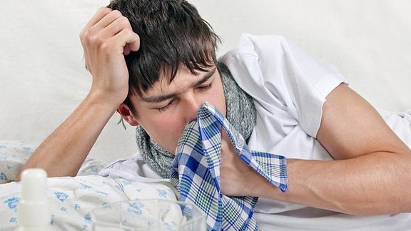 Ein junger Mann liegt im Bett und hält sich ein Taschentuch vor den Mund