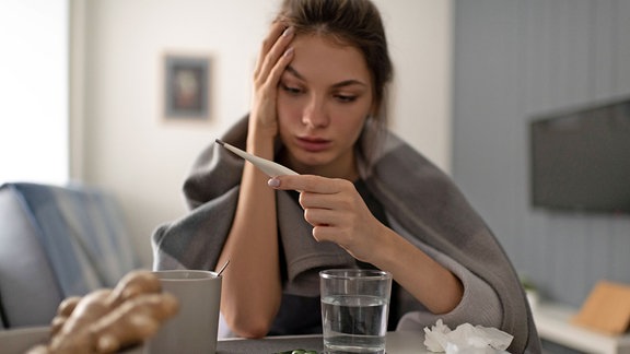 Junge Frau mit einer Erkältung schaut auf ein Fieberthermometer.