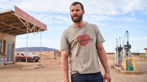 Ein Mann steht an einer Tankstelle in einer trockenen Wüstenlandschaft.