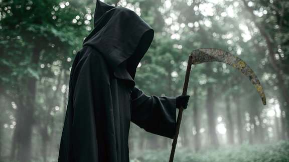 Sensenmann - Eine Person im schwarzen kaputzenumhang und mit Sense steht im Wald.