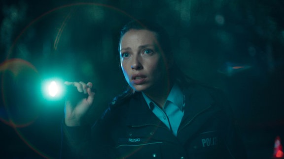 Eine Frau in Polizeiuniform und mit zusammengebundenen Haaren leuchtet mit einer Taschenlampe durch die Dunkelheit.