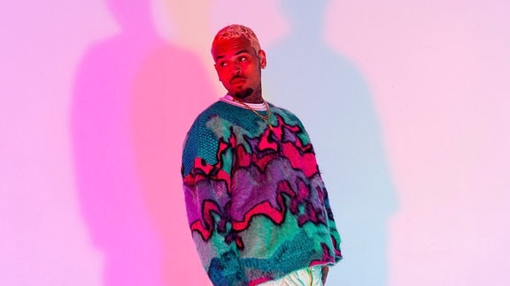Der Künstler Chris Brown steht in einem bunten Pullover vor einer Wand