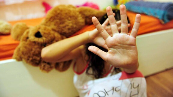 Ein junges Mädchen, vor einem Bett auf dem Boden hockend, hält schützend die Hände vor sich
