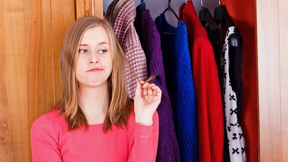 Eine junge Frau steht unschlüssig vor einem offenen Kleiderschrank