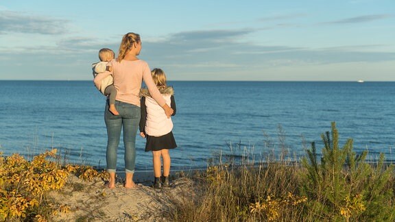 Eine Mutter steht mit ihren zwei kleinen Kindern in Schleswig-Holstein am Ufer der Ostsee und schaut aufs Meer
