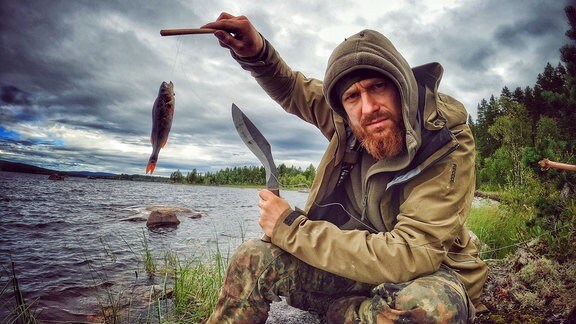 Ein Mann schaut in die Kamera und hält einen Fisch an einer Angelschnur hoch.
