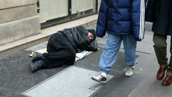 Ein obdachloser Mann liegt auf der Straße, Passanten gehen daran vorbei