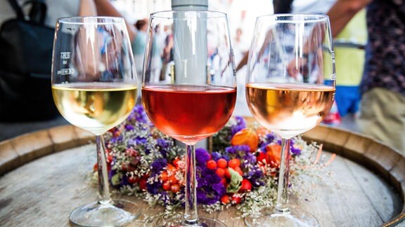 Glas Weisswein, Rotwein und Rose, von links nach rechts, auf einem Holzfass stehend mit Blumenschmuck dahinter