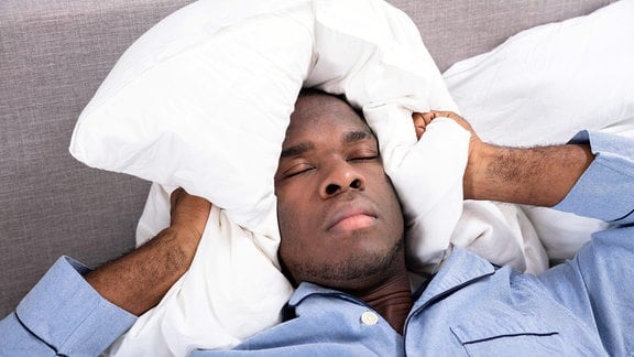 Ein Mann liegt mit geschlossenen Augen im Bett und hält sich mit einem Kissen die Ohren zu. 