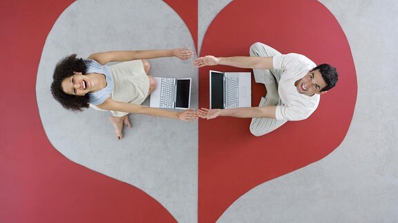 Eine Frau und ein Mann sitzen auf einem Teppich mit Herz-Muster vor ihren Laptops