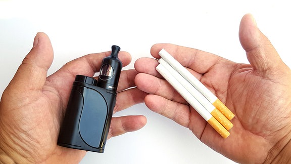 Mann hält in seiner linken Hand eine E-Zigarette und in seiner rechten Hand drei herkömmliche Tabakzigaretten.