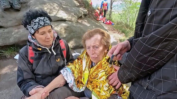 Eine verletzte Frau in einer Wärmfolie und ihren Begleitern. Im Hintergrund die Gruppe der Bergwacht.