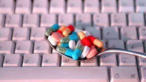 Ein Löffel mit verschiedenfarbigen Medikamenten wird über eine Computertastatur gehalten. 