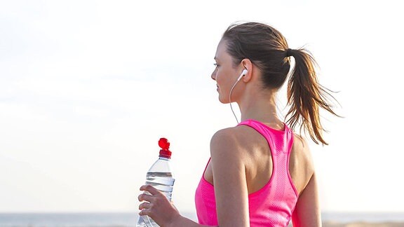 Eine junge blonde Frau in Sportkleidung und mit Kopfhörern steht mit einer Getränkeflasche in der Hand an einem Strand