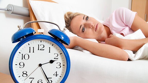 Eine junge Frau liegt schlaflos im Bett. Auf ihrem Nachttisch steht ein Wecker, der die Zeit 2:25 Uhr anzeigt.
