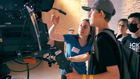 Eine junge Fau erklärt Jugendlichen eine TV-Kamera.