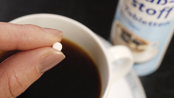 Finger halten kleine weißeTablette über Kaffeetasse, daneben Dose mit Aufschrift "Süßstoff"