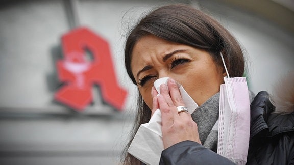 Junge Frau, mit am Ohr hängendem Mundschutz, putzt sich die Nase