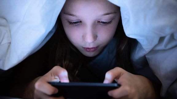 Ein Kind spielt unter der Bettdecke mit einem Smartphone