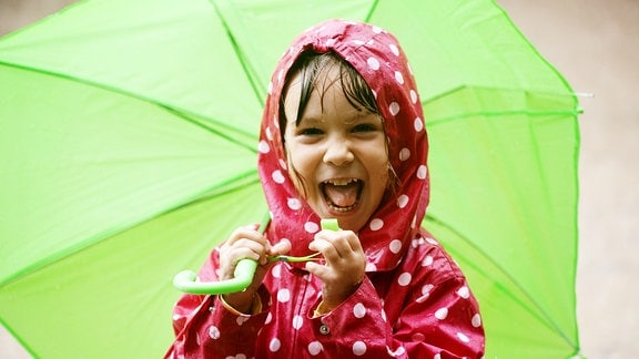 Lachendes Kind in rotem Anorak mit weißen Punkter und grünem Regenschirm im Regen.